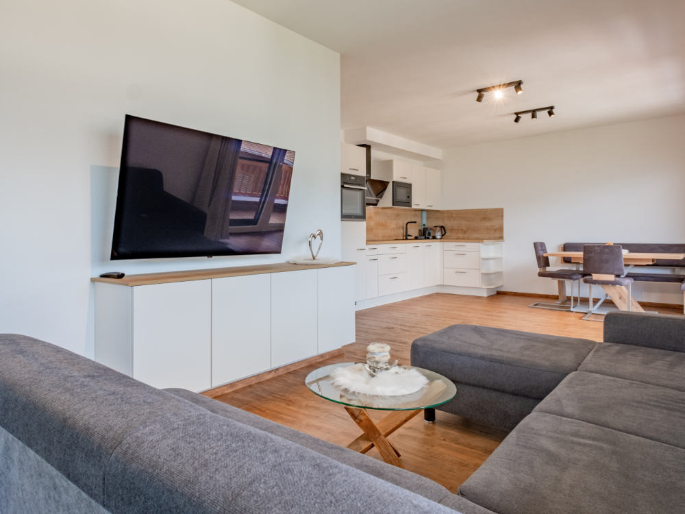 Wohnraum der Ferienwohnung Hoferlitzlhubgut mit gemütlichem Sofa, großem Fernseher und offener Küche im Hintergrund.