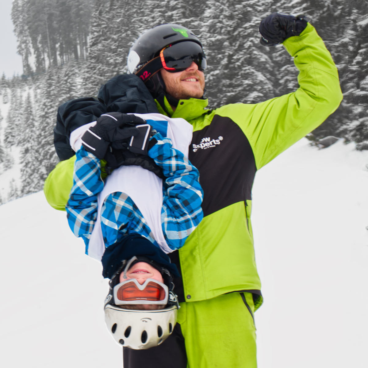 Snow Experts Skilehrer hält ein Skikurs-Kind kopfüber, beide lachen und haben sichtlich Spaß im Skigebiet KitzSki.